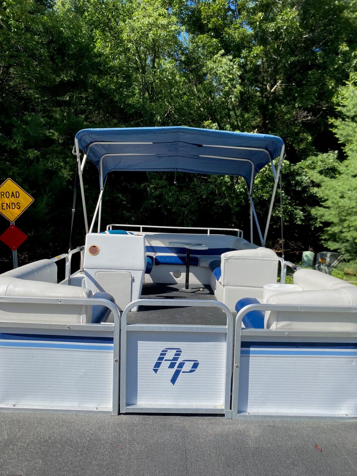 White and blue HP pontoon boat with canopy top parked on street, trees in background
