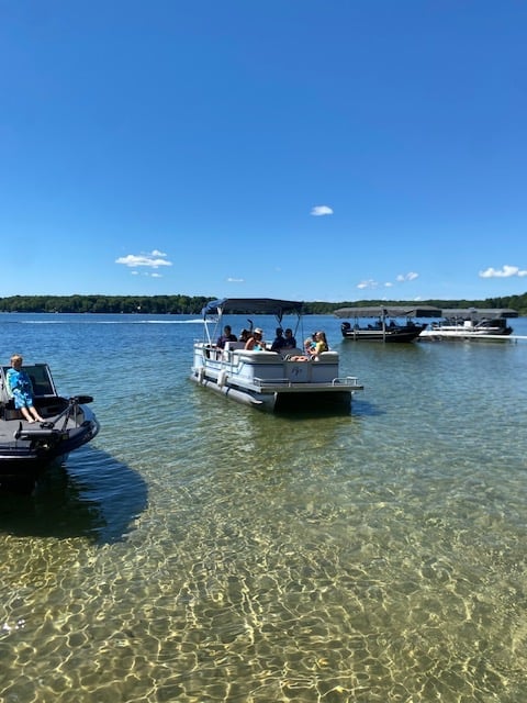 Pontoon boat with passengers in clear blue water, with additional boats and forested shoreline in background under blue sky