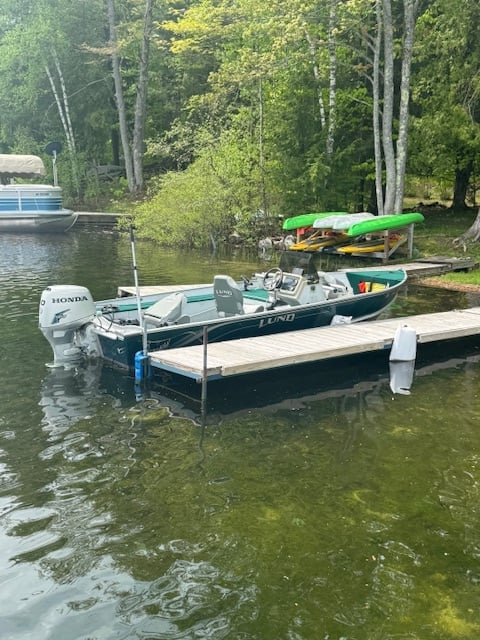 Blue fishing boat with Honda motor docked at wooden pier on calm lake surrounded by tall trees and forest