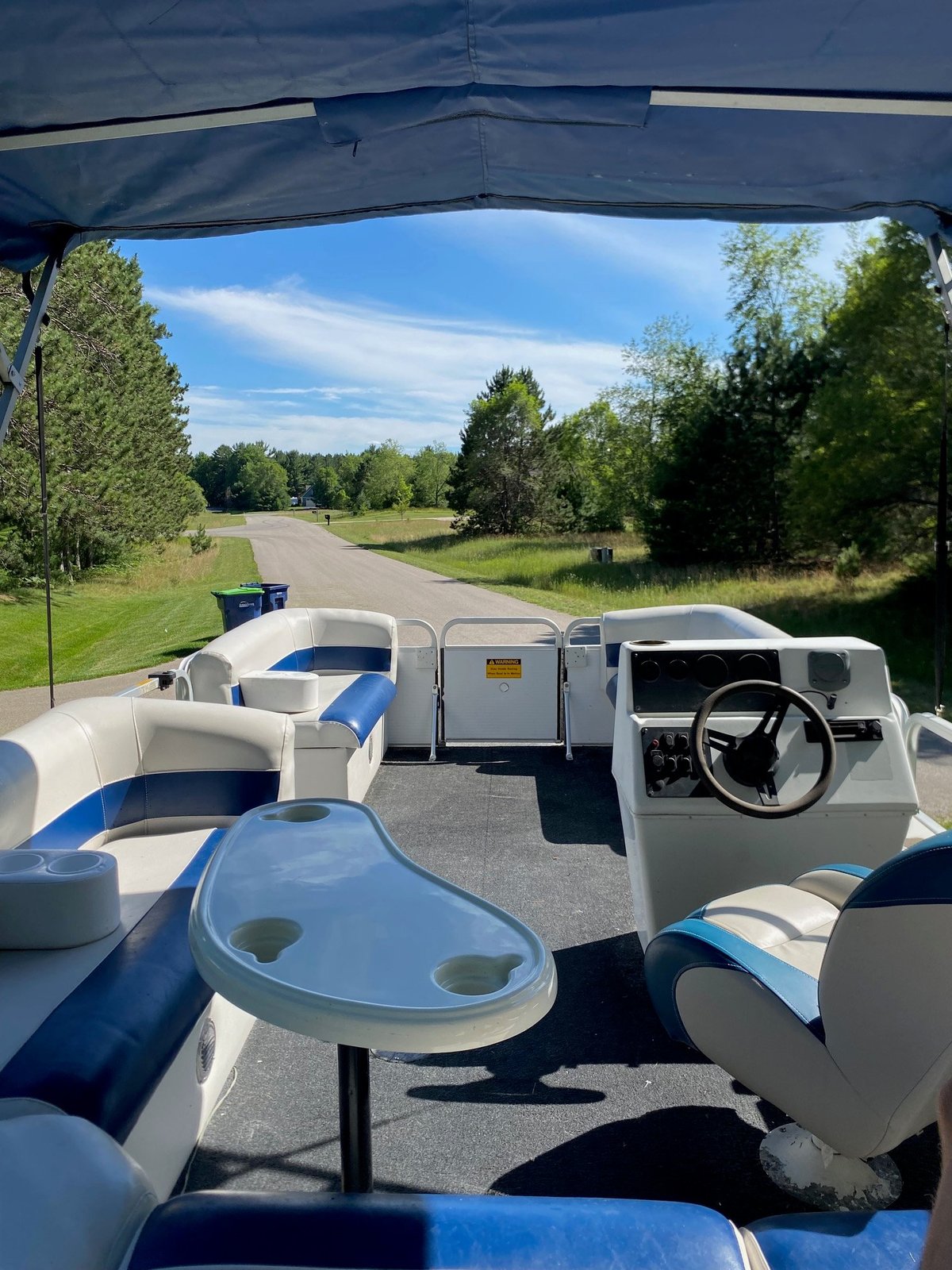 Interior view of a blue and white pontoon boat on a rural road with trees and clear sky visible from under the canopy