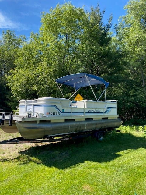 Blue and white pontoon boat with canopy on grass near trees under clear sky