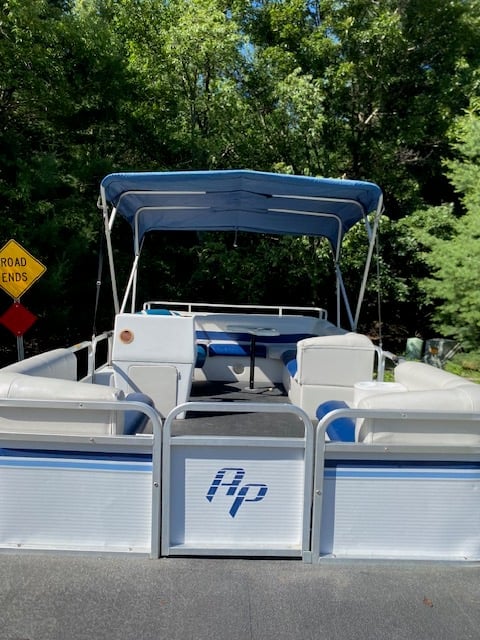 White and blue pontoon boat with blue canopy top parked on road with dense green trees in background