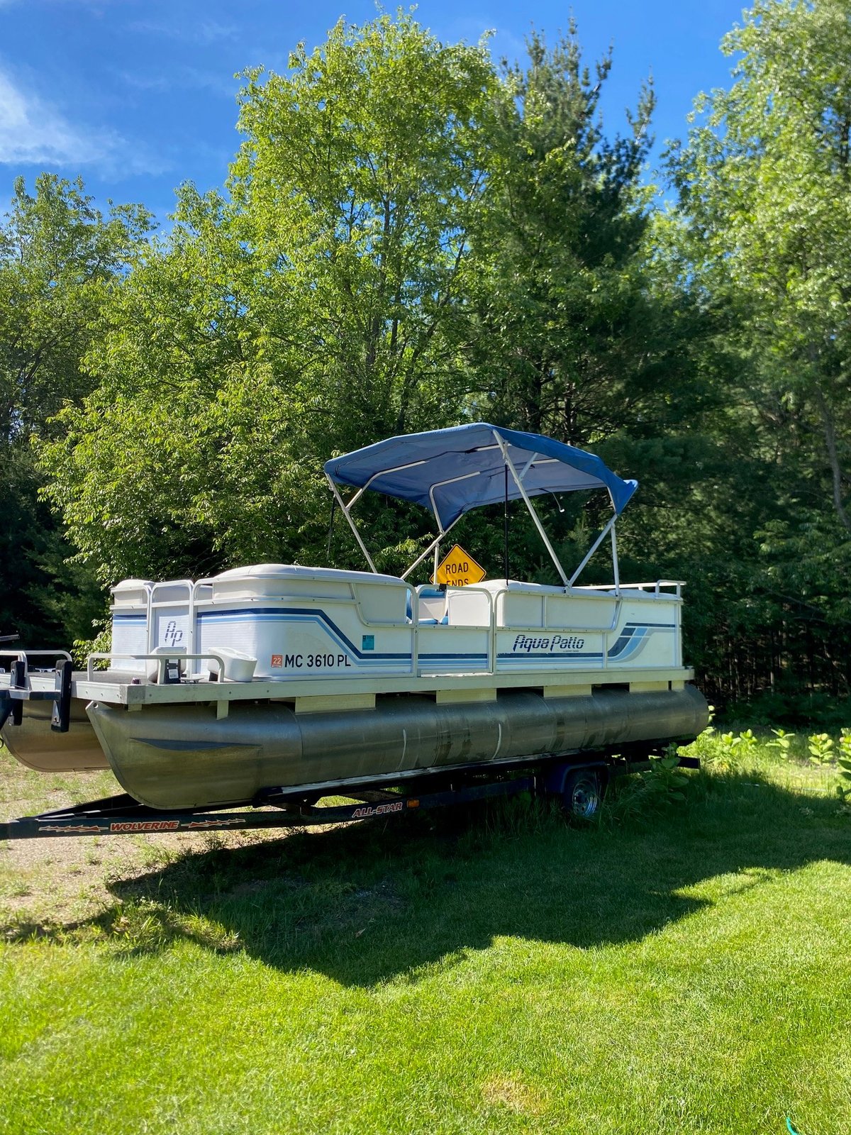 White and blue pontoon boat with blue canopy parked on grass near green trees on sunny day