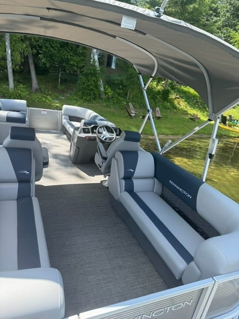 Interior of a pontoon boat with gray and white seating, steering wheel, and shaded canopy, docked near trees along a waterway