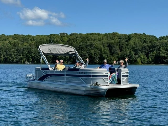 A pontoon boat with passengers on blue water, forested shoreline and clear sky in background